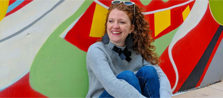 Jamie sitting at mural in Crosstown Concourse with SALT Lorna Sunglasses on and Nickel and Suede earrings and JCrew Factory bow sweater