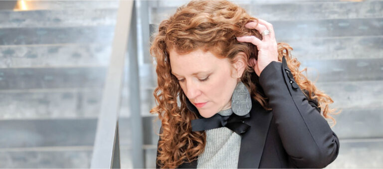 Jamie is looking down and holding hair back. She is wearing the J Crew tie neck sweater, a black tuxedo blazer and Nickel and Suede Black cork earrings.